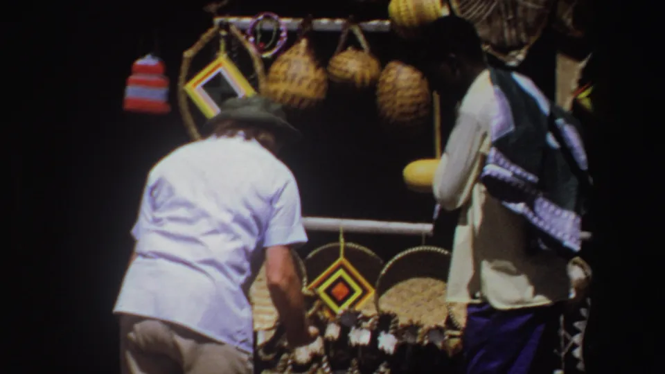 Adult men browse a South African market display with baskets, hanging craftwork, and a yellow object in bright daylight.