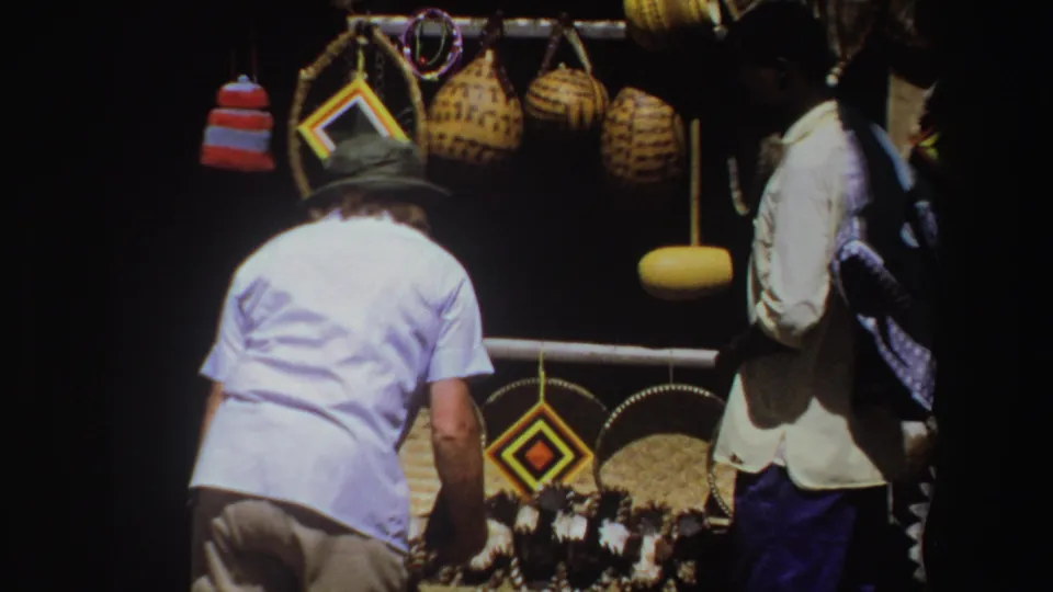 South African 8mm home-movie frame of adult men beside a market stall filled with woven baskets and patterned crafts.