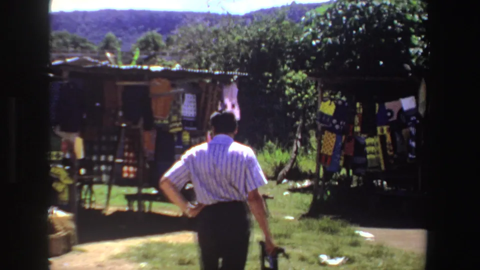 Wide 1969 South Africa frame showing a man facing outdoor craft displays, grassy ground, trees, and distant hills.