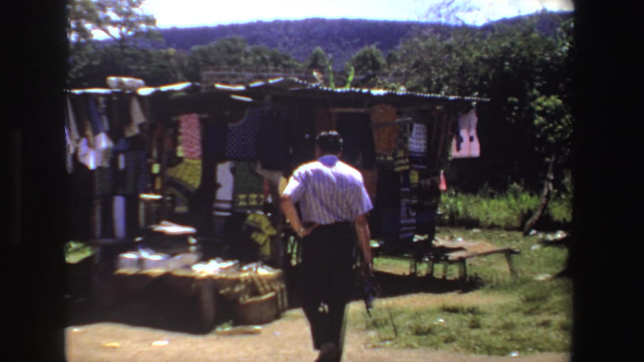 The man continues toward the South African rural clothing stall, with bright garments under a corrugated roof.