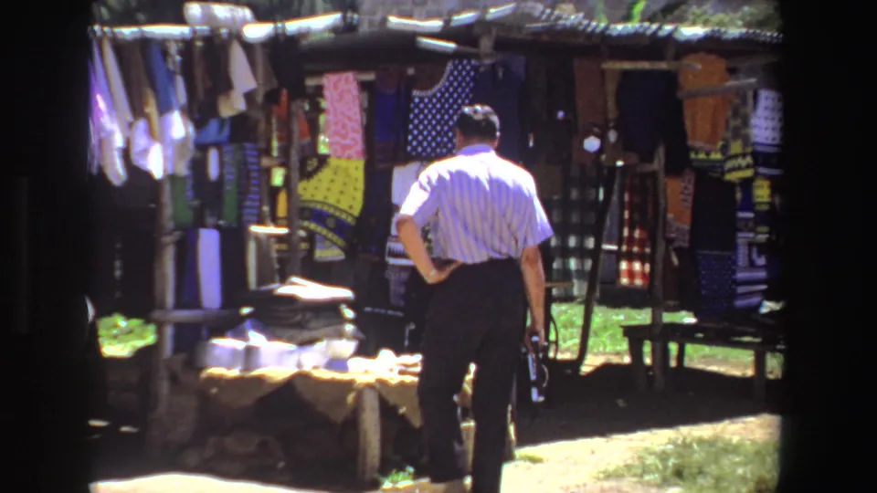 A closer 1969 view shows the man pausing near colorful clothing and fabric displayed at the rural stall.