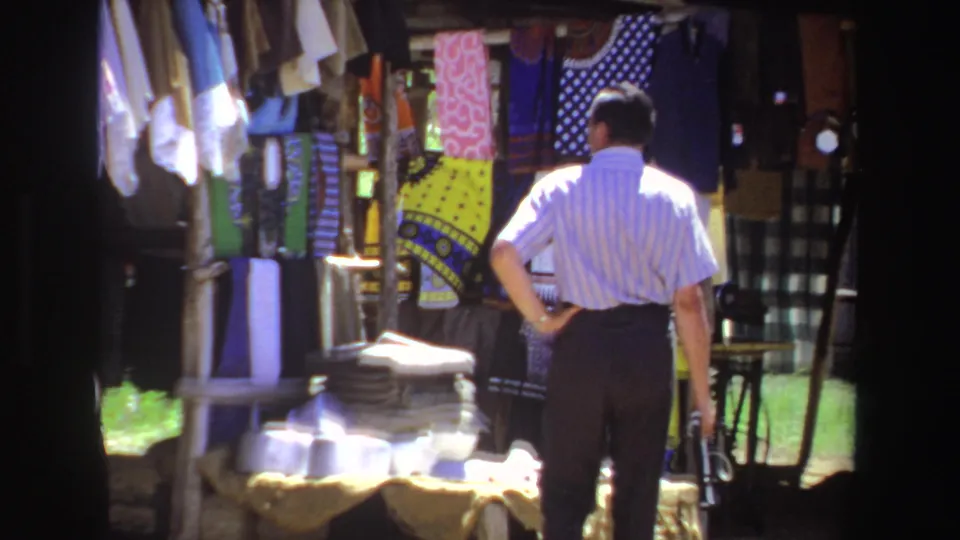The final frame shows the man beside hanging garments and a table of wares at the South African outdoor stall.