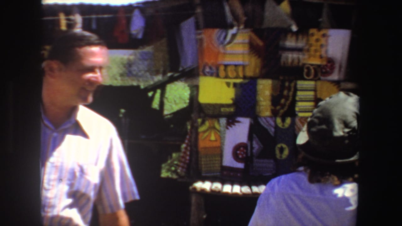 Man in striped shirt and woman in hat stand near colorful textiles at an outdoor stall.