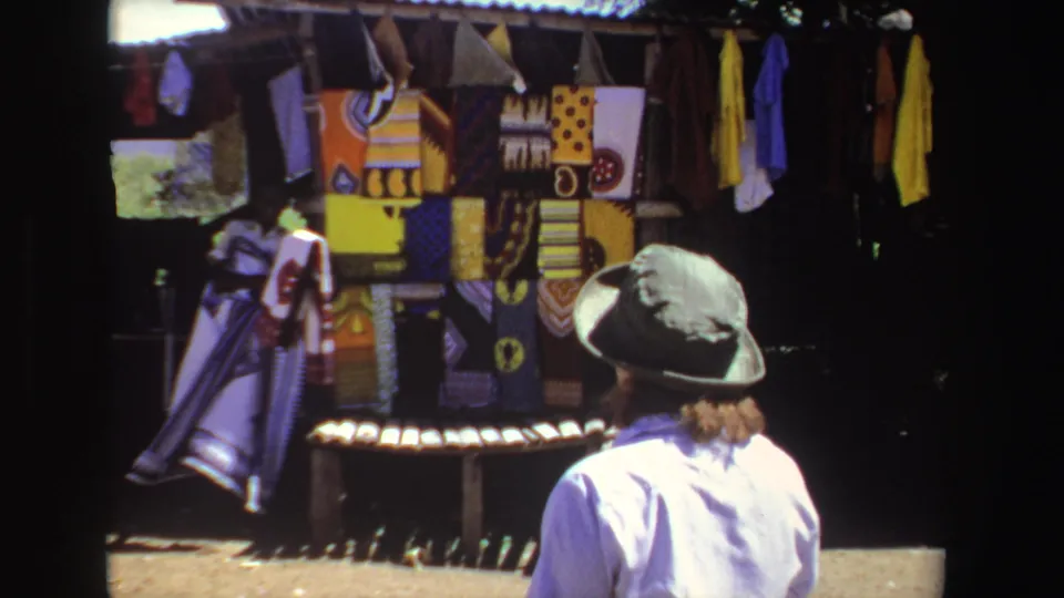 The woman is seen from behind as she looks toward a rack of colorful textiles at a shaded South African outdoor market stall.