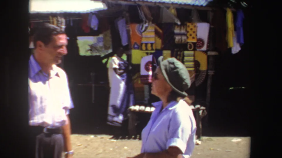 A man and woman converse beside a display of vivid patterned textiles at a South African outdoor market, captured in 1969 home-movie footage.