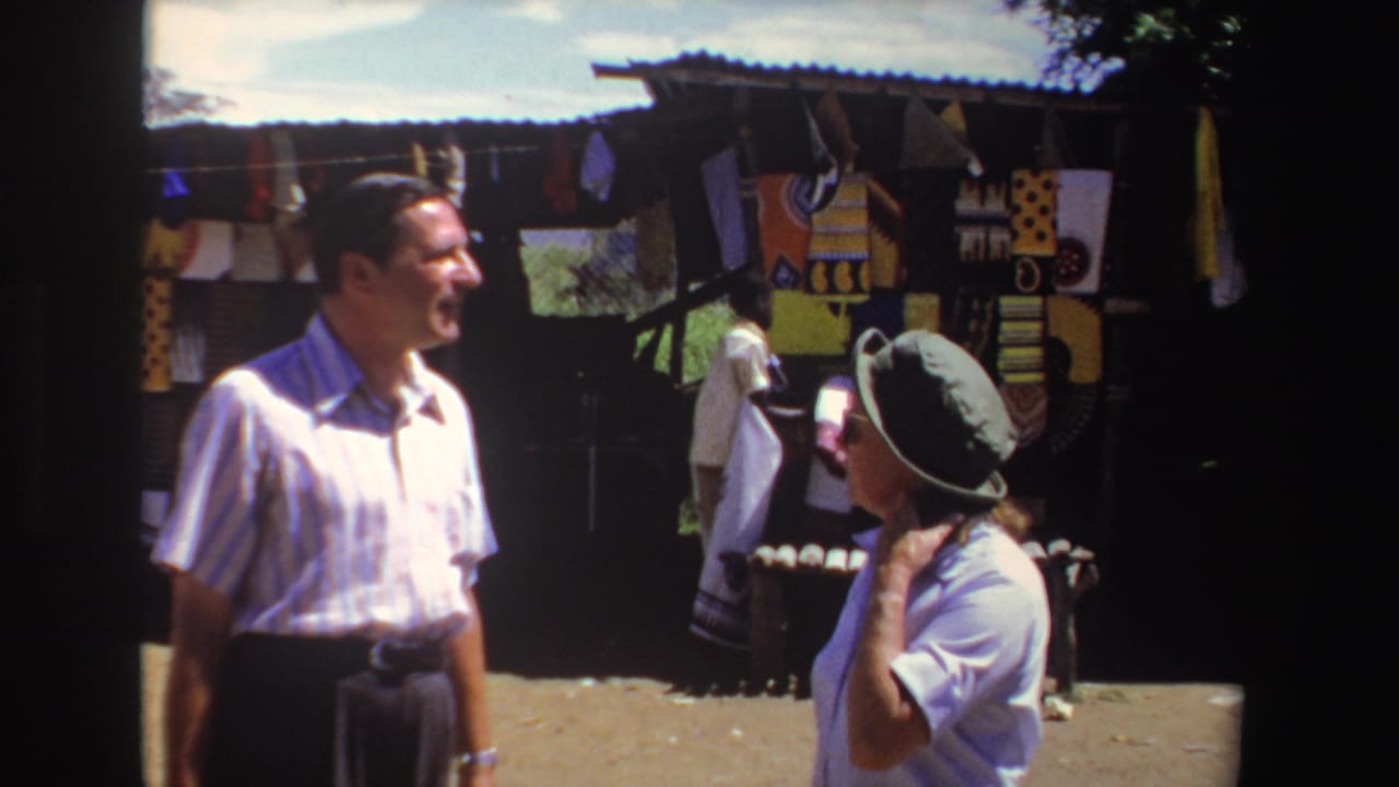 In 1969 South Africa footage, two adult tourists stand in bright sun near a market stall displaying patterned textiles.