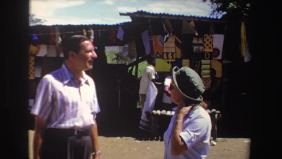 In 1969 South Africa footage, two adult tourists stand in bright sun near a market stall displaying patterned textiles.