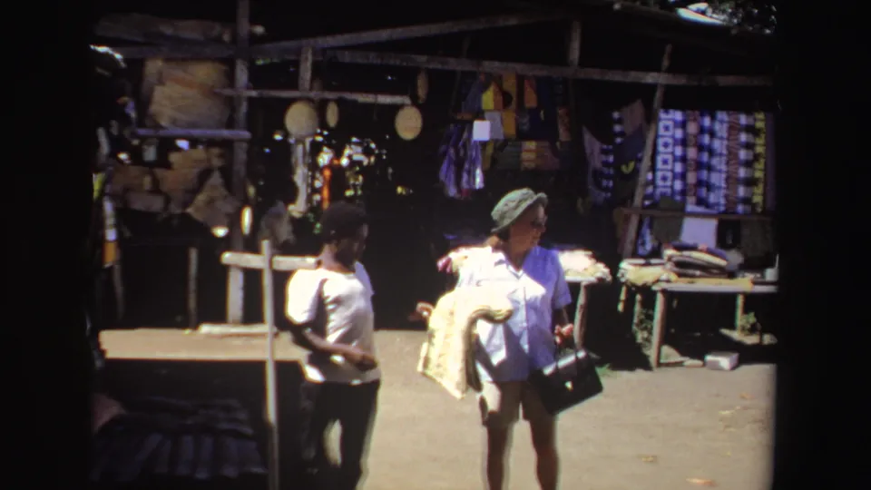 The woman tourist walks with a folded patterned textile near a merchant and souvenir displays in the 1969 marketplace scene.