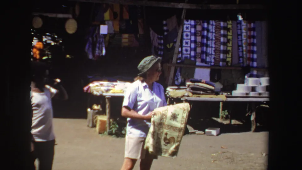 A woman tourist in South Africa holds a patterned textile in front of hanging fabrics and folded goods at an outdoor market.