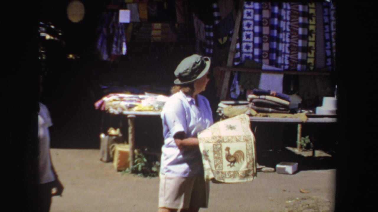 In 1969 South Africa home-movie footage, a tourist in a hat examines a rooster-pattern towel beside hanging textiles.