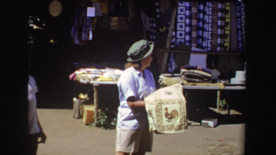 In 1969 South Africa home-movie footage, a tourist in a hat examines a rooster-pattern towel beside hanging textiles.