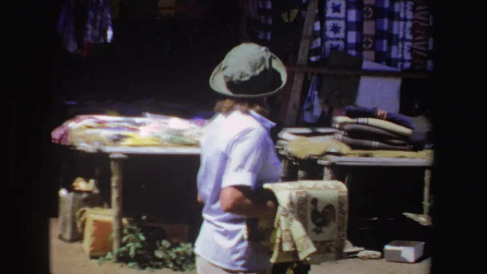 A tourist in a light shirt and hat stands near a South African market stall with folded textiles and a rooster towel.