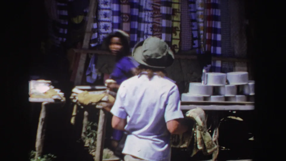 At the market stall, the tourist holds the rooster towel while another person appears among textiles and stacked bowls.