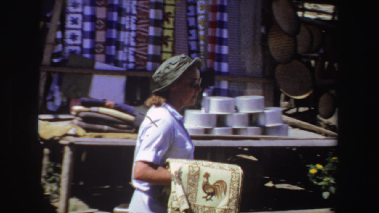 Woman in a green sun hat carries a patterned cloth past market stalls.