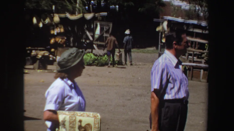 The final frame shows the woman and a man passing through the South African market, with craft goods, baskets, and shaded stalls behind them.