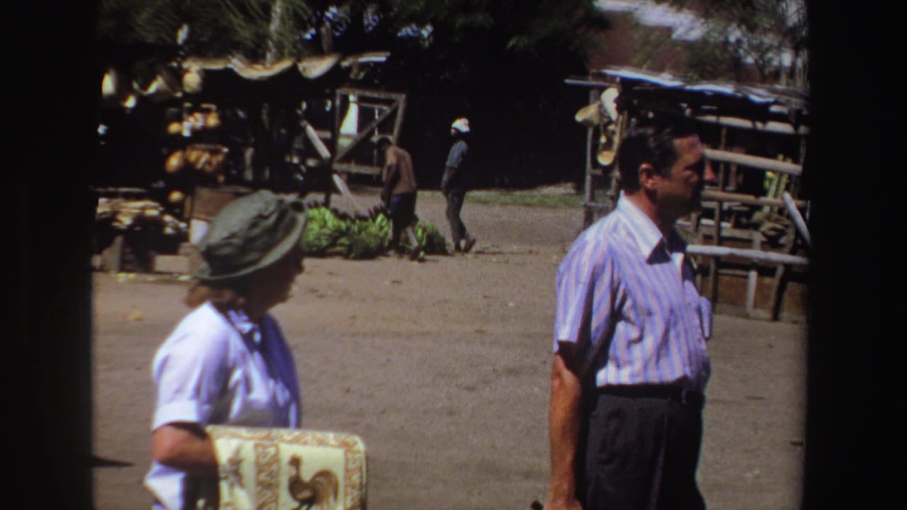 In 1969 South Africa home-movie footage, a woman with a hat and decorative box stands near a man in a blue striped shirt by a rustic market stall.