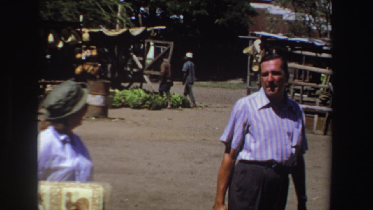 Man in a striped shirt faces the camera while a woman in a hat passes at the left edge.