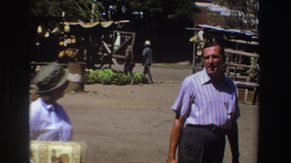 The man in a blue striped short-sleeve shirt looks toward the camera as the woman in a hat moves nearby in the South African market setting.