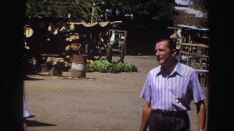 A man in a blue striped shirt is framed closer in the 1969 South Africa home movie, with a rustic stall and shaded outdoor area behind him.