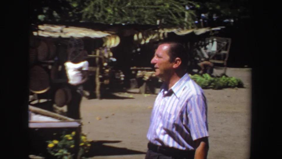 The man appears in profile beside a shaded rural market stall, with hanging produce and wooden fixtures visible in the 8mm South Africa footage.
