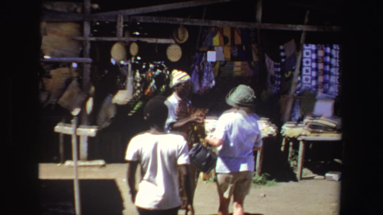 1969 South Africa 8mm frame showing adults beside a shaded outdoor textile stall with hanging fabrics and market goods.