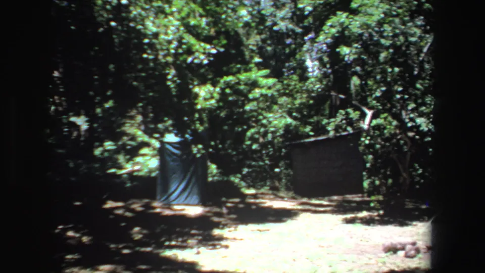 1969 South Africa 8mm frame showing a rustic campsite with tarp shelter, dark tent wall, trees, and stones on the ground.