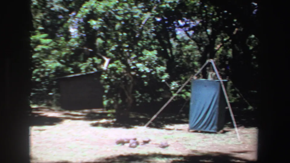 1969 South Africa 8mm frame of a forest campsite with a blue tarp shelter supported by poles and surrounded by trees.