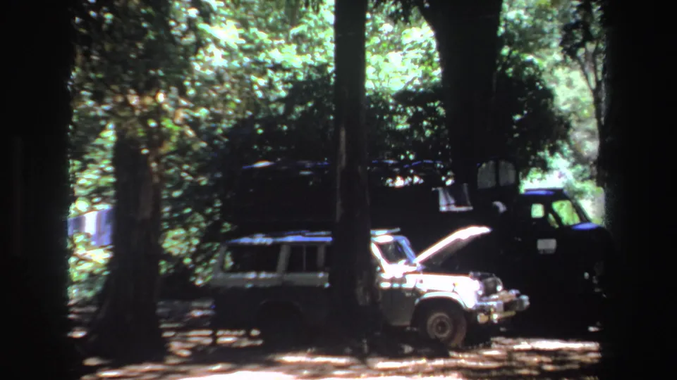 Closer 8mm view of the white car's raised hood and a dark utility truck parked under trees in South Africa.