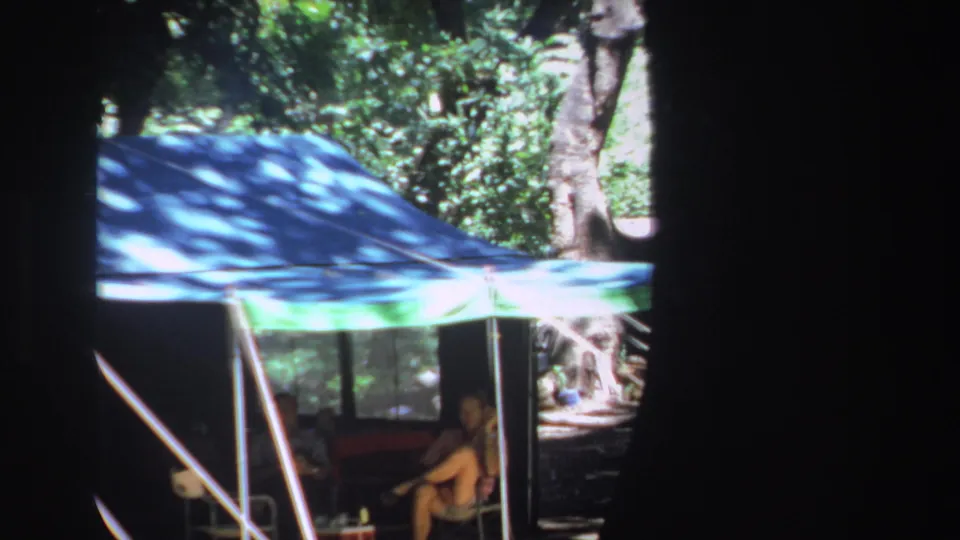 1969 South Africa campsite frame showing a blue green tarp shelter among trees, with a seated adult figure visible underneath.