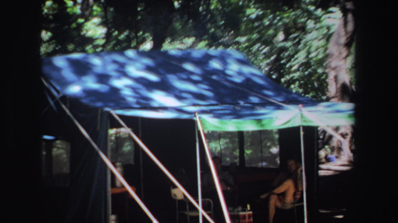 1969 South Africa 8mm frame showing a man in shorts seated beneath a shaded blue and green camping canopy among trees.