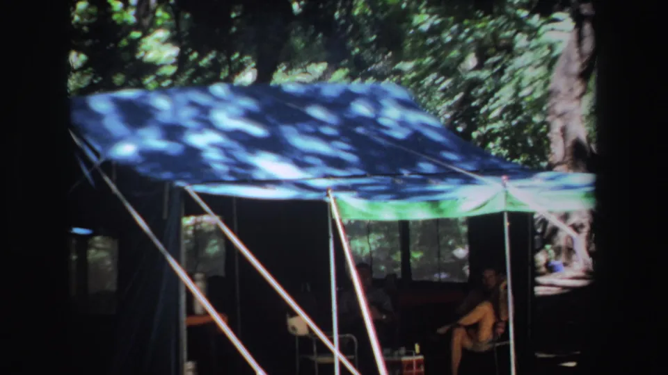 1969 South Africa 8mm frame showing a man in shorts seated beneath a shaded blue and green camping canopy among trees.