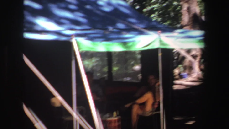 1969 South Africa frame showing the man still seated under the camping shelter, with angled poles and trees in the background.
