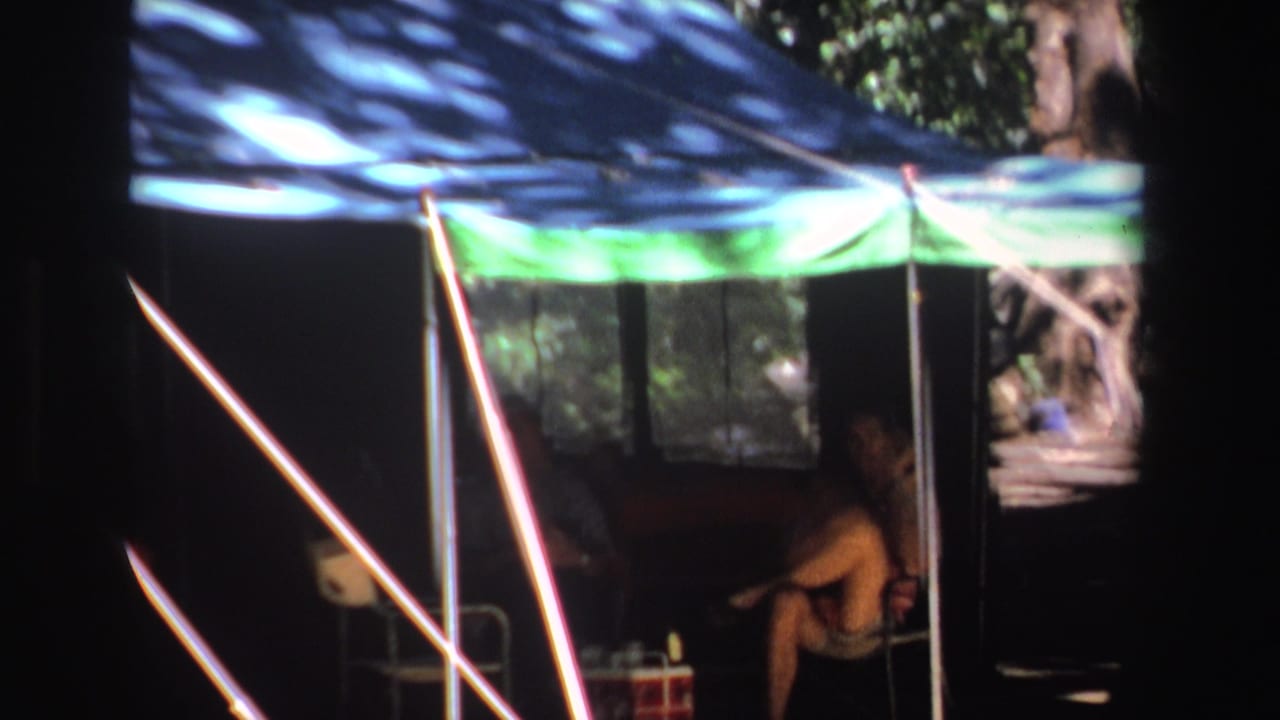 Person seated inside a shaded tent under a blue and green canopy.