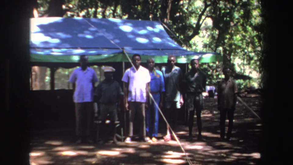 1969 South Africa 8mm frame showing eight men standing in a line near a green tent under trees with dappled sunlight.