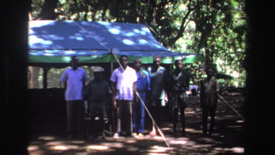 1969 South Africa 8mm frame of men standing near a green tent in a wooded area, with strong shade and sun patches across the scene.