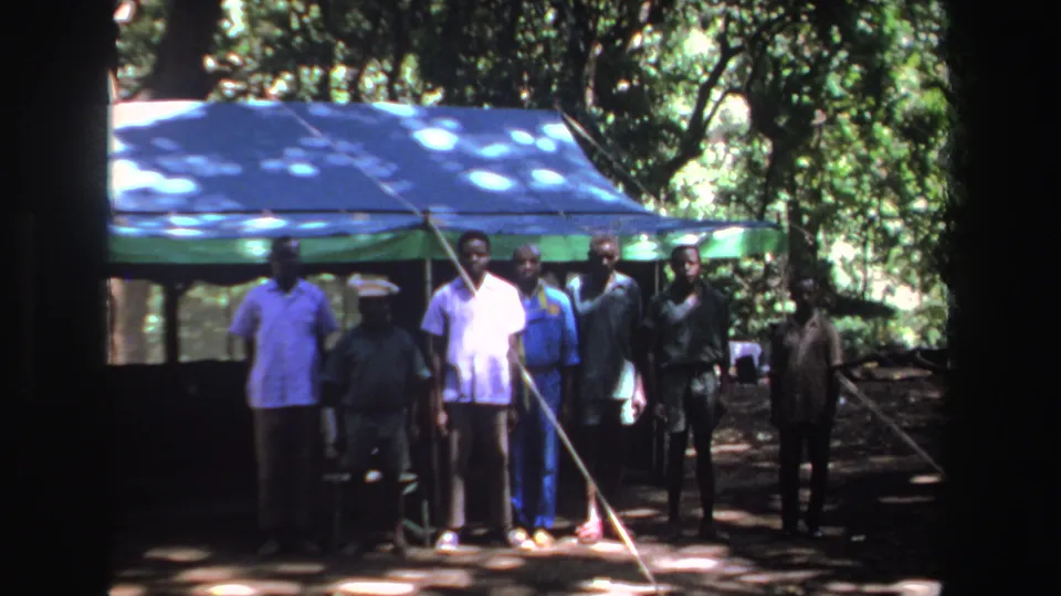 1969 South Africa 8mm still of seven Black men standing before a blue and green tent beneath trees.