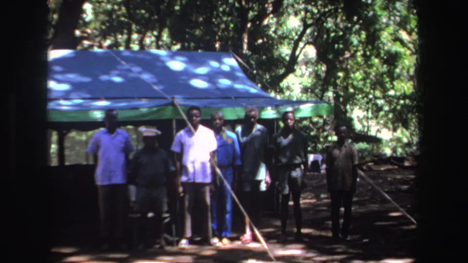 1969 South Africa frame with several Black men standing under trees beside a blue tarp shelter.