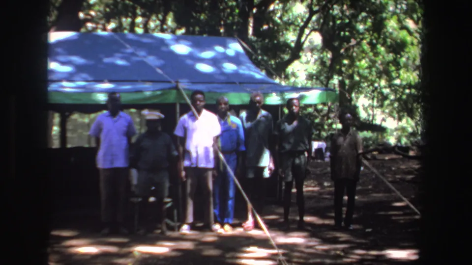 Black men stand in front of a blue tarp shelter in wooded 1969 South Africa 8mm footage.