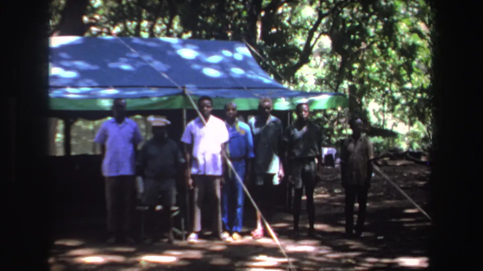 Several men stand beneath trees by a tarp shelter in South Africa, supported by 1969 8mm home-movie metadata.