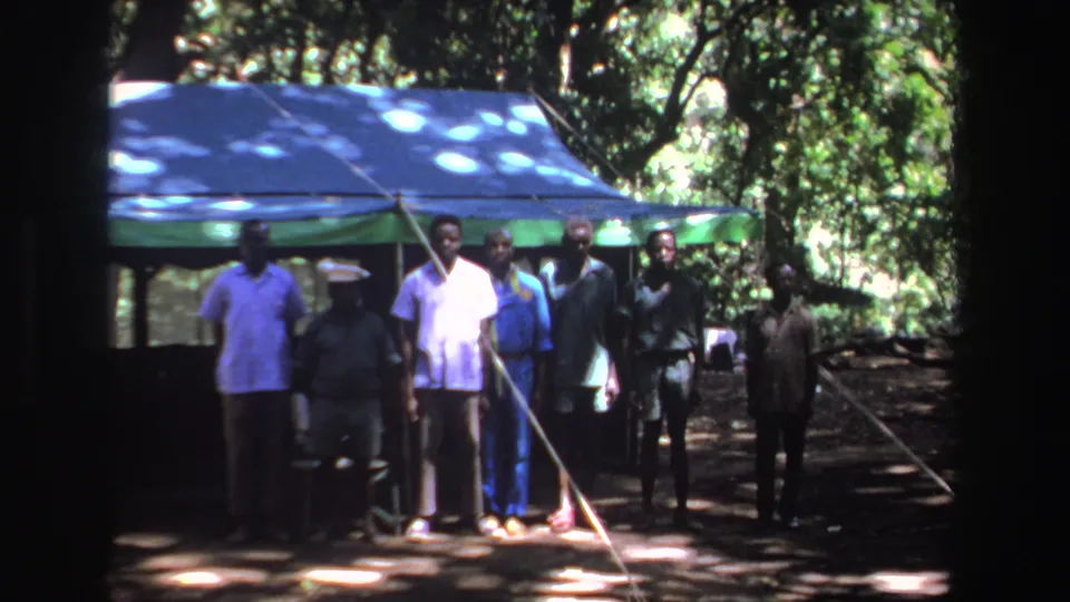 A group of men faces the camera beside a blue and green tarp shelter in wooded South Africa, from 1969 8mm footage.