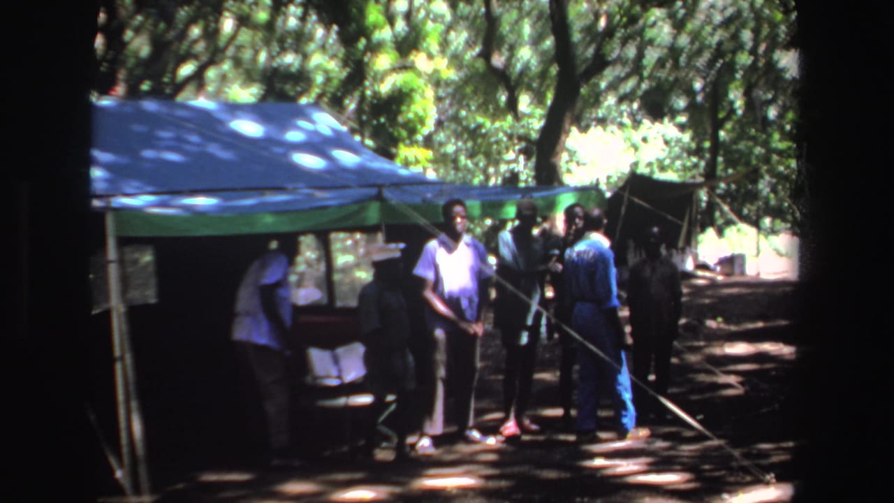 Men stand and lean near a tent in dappled shade.