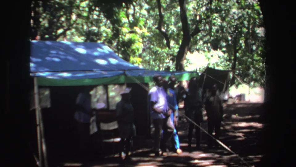Several men cluster beside the tent entrance under trees, with strong shadows and soft 8mm color in the 1969 South Africa footage.