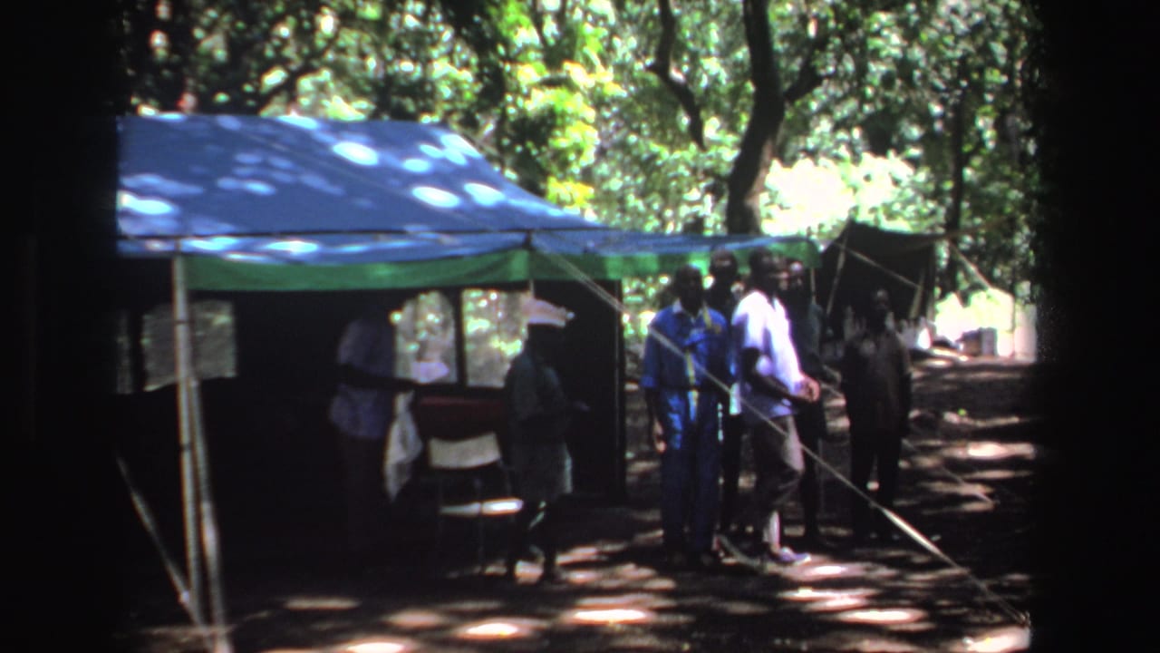 People stand near a blue and green canopy tent in a wooded area.