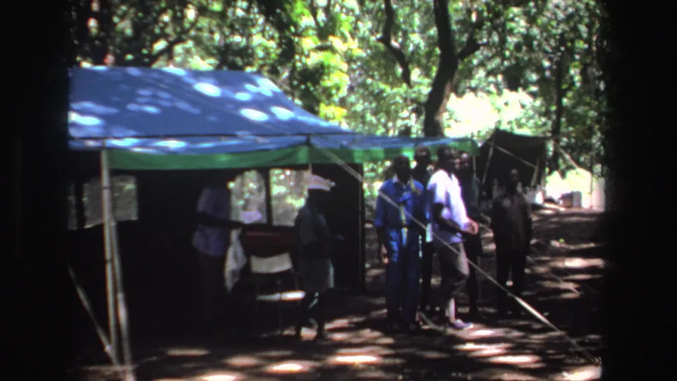 1969 South Africa 8mm frame showing adults gathered beside a blue and green canopy tent under trees.