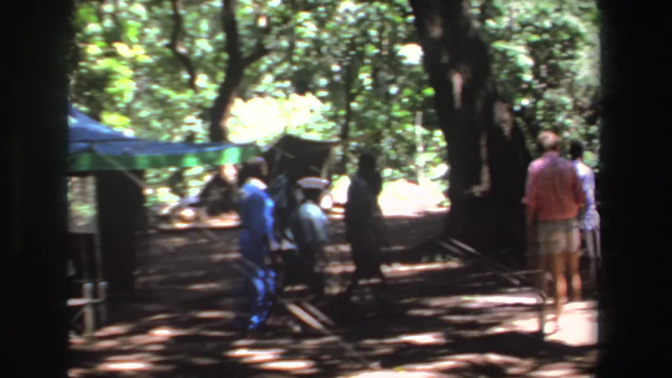 A home-movie view of people moving through a wooded gathering area with tent ropes and filtered sunlight.
