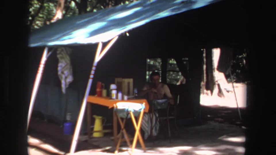 1969 South Africa campsite frame with a seated adult under a tarp beside folding furniture and food or drink containers.