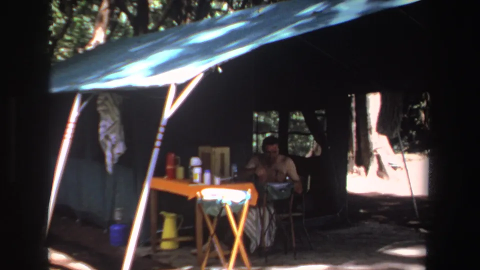 A home-movie view of the shaded campsite shelter, with a seated adult and picnic items under dappled forest light.