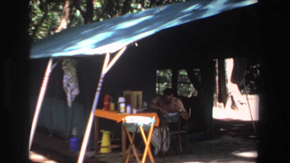 1969 South Africa 8mm frame showing a man at a camp table beneath a blue tarp, with thermoses and hanging clothes nearby.