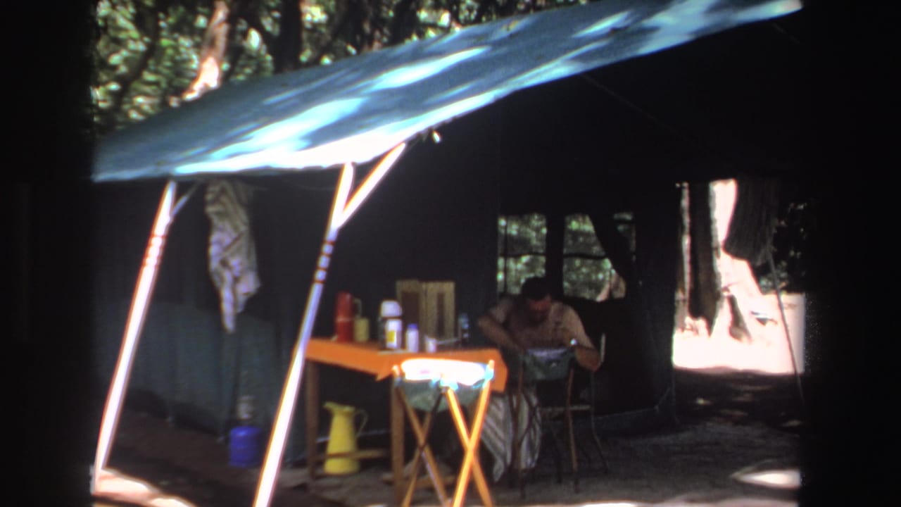 1969 South Africa home-movie frame of a shaded campsite, with a man seated near a folding table holding bottles and gear.