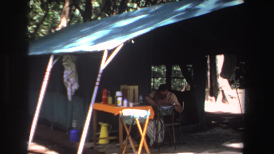 1969 South Africa home-movie frame of a shaded campsite, with a man seated near a folding table holding bottles and gear.